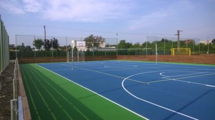 Kolorowe boisko sportowe z koszem i bramką, otoczone ogrodzeniem.