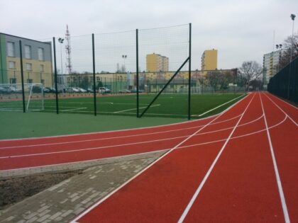 Bieżnia i boisko sportowe otoczone ogrodzeniem, w tle bloki mieszkalne